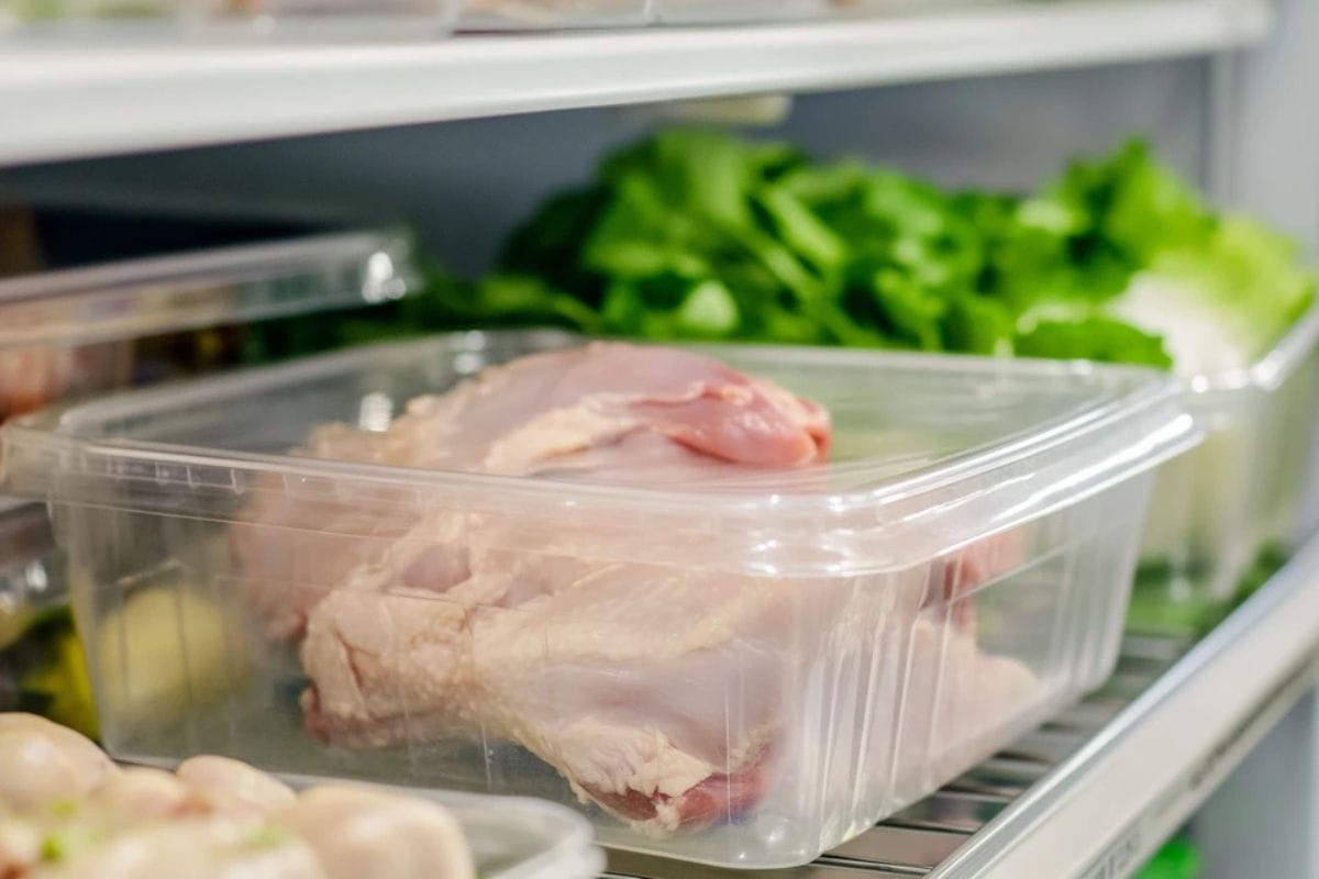 A sealed container with raw chicken defrosting on the bottom shelf of a refrigerator.